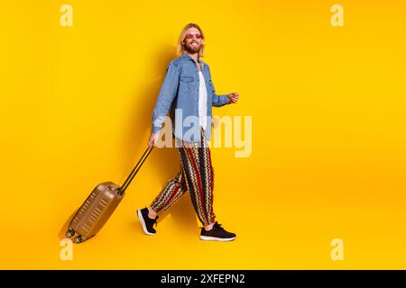 Foto a grandezza naturale di un viaggiatore di valigia da giovane, indossa un abito di jeans alla moda isolato su sfondo giallo Foto Stock