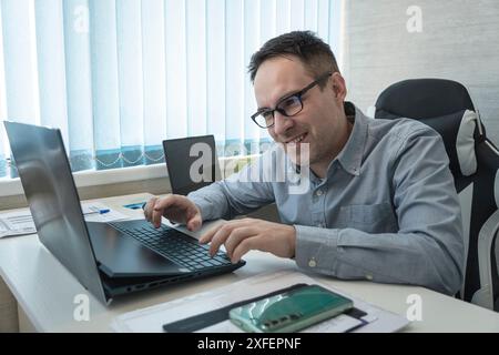 Elegante uomo d'affari che analizza i dati in ufficio. un giovane lavoratore con una camicia blu e occhiali sorride in ufficio. Foto Stock