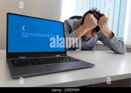 Visualizzazione di una schermata blu di errore sul notebook sullo sfondo di un uomo arrabbiato che stringe la testa Foto Stock