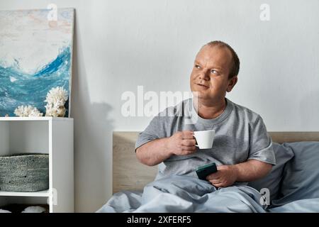 Un uomo con inclusività siede a letto, sorseggiando una tazza di caffè e guardando attento. Foto Stock