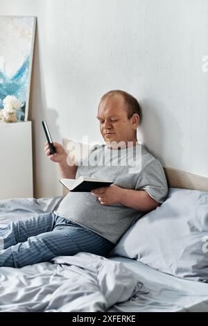 Un uomo con l'inclusività si siede a letto, legge un libro e tiene il telefono. Foto Stock