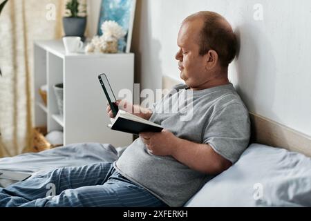 Un uomo con l'inclusività si siede a letto, guardando il suo telefono mentre tiene un libro. Foto Stock