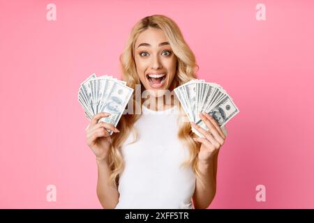 Donna eccitata che tiene in mano contanti su sfondo rosa, esclamando Foto Stock