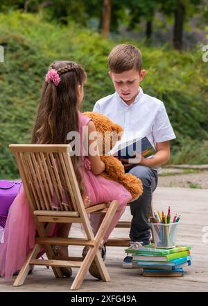 Una bambina con i capelli lunghi si siede con la schiena, tiene un orsacchiotto tra le mani e ascolta attentamente mentre un ragazzo legge un libro. Nelle vicinanze c'è una scuola Foto Stock