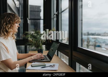 Donna d'affari concentrata con capelli ricci che lavora su un computer portatile mentre è seduta vicino alla finestra di casa Foto Stock