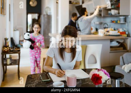 Donna matura che scrive nel libro mentre siede a tavola nel soggiorno di casa Foto Stock