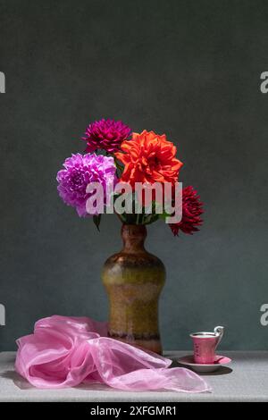 Natura morta con fiori. Dahlias rosa e rosse in vaso scuro, tessuto rosa, piccola tazza rosa, sfondo scuro Foto Stock