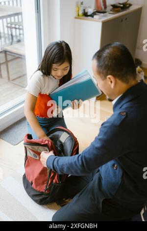 Vista ad alto angolo dello zaino della figlia con il padre inginocchiato sul pavimento di casa Foto Stock