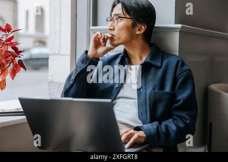 Un uomo d'affari premuroso seduto con un computer portatile sul davanzale della finestra in ufficio Foto Stock