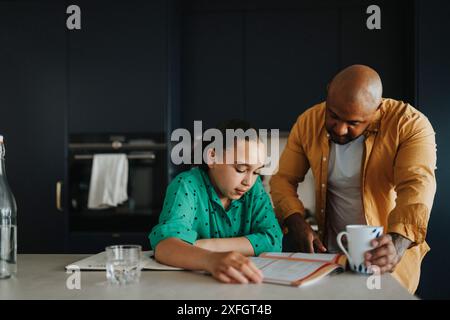 Padre che aiuta la figlia negli studi mentre tiene una tazza di caffè a casa Foto Stock