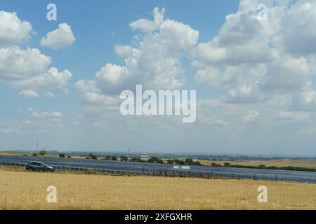 Una singola auto percorre una strada sterrata attraverso un campo di pannelli solari in un paesaggio rurale sotto un cielo azzurro e limpido, pieno di soffici nuvole bianche. Foto Stock