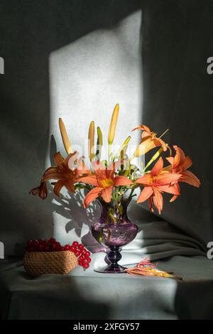Un bouquet di gigli d'arancia, un cesto con bacche rosse accanto. Un raggio di luce cade sui fiori. Natura morta Foto Stock
