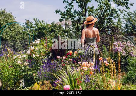 Donna giardiniera in cappello di paglia che cammina nel suo giardino estivo ammirando fiori di rose. Controllo degli impianti Foto Stock