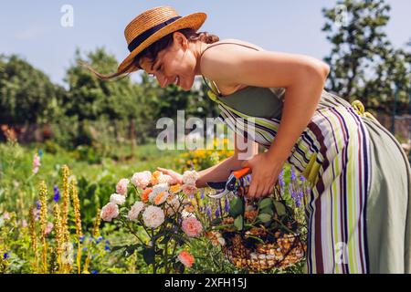 Ritratto di una donna che ha trascorso fiori di rosa nel giardino estivo. Il giardiniere tiene il cesto con fiori e potatrice selvaggi. Prendersi cura delle piante Foto Stock