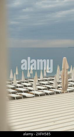 Una tranquilla spiaggia di Nizza con lettini e ombrelloni chiusi disposti in modo ordinato, affacciata sul tranquillo Mar Mediterraneo sotto un cielo nuvoloso. Foto Stock