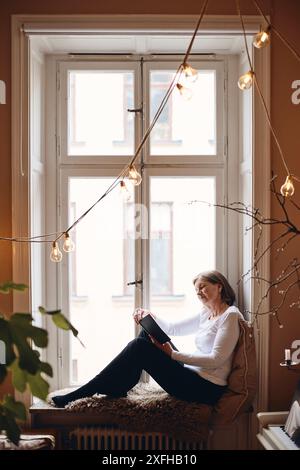 Donna anziana che legge un libro seduto sul davanzale della finestra a casa Foto Stock