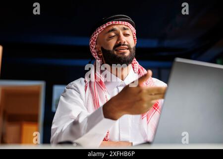 Giovane uomo d'affari che rappresenta il successo nell'era digitale, indossa abbigliamento arabo e utilizza un computer portatile digitale. Un musulmano che parla con sicurezza sul suo personal computer alla sua scrivania di casa. Foto Stock