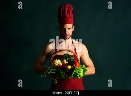 Il bellissimo chef maschile in grembiule tiene il cestino di vimini con verdure fresche per il ristorante. Consegna di cibo. Preparazione di alimenti sani, cottura. Servizio di ristorazione. C Foto Stock