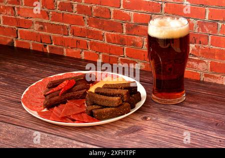 Un alto bicchiere di birra scura e un piatto con un assortimento di spuntini di carne con crostini e formaggio su un tavolo di legno. Primo piano. Foto Stock