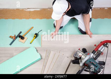 lavoratore maschio che posa pavimenti in vinile o laminato. utensili come la sega a tazza, il laminatoio misura le piastrelle in laminato grigio sull'isolamento acustico del gradino. profe. fai da te Foto Stock