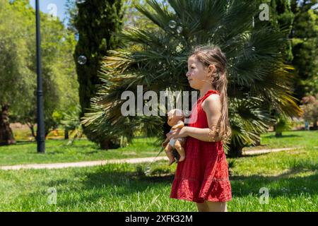 Un'affascinante ragazza vestita con un vivace abito rosso tiene con amore la bambola mentre si gode una giornata di sole nel parco. Intorno a lei, le bolle di sapone fluttuano nella A. Foto Stock