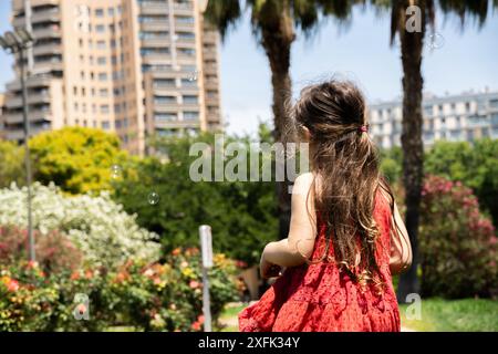 Una ragazza vestita con un affascinante vestito rosso viene vista da dietro, circondata da bolle di sapone che fluttuano nell'aria in un parco verde. Le palme sono alte in t Foto Stock
