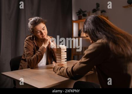 Due donne stanno giocando a un gioco a blocchi impilabili su un tavolo di legno in una stanza accogliente con illuminazione calda. Una donna sta considerando attentamente la sua mossa mentre il Foto Stock