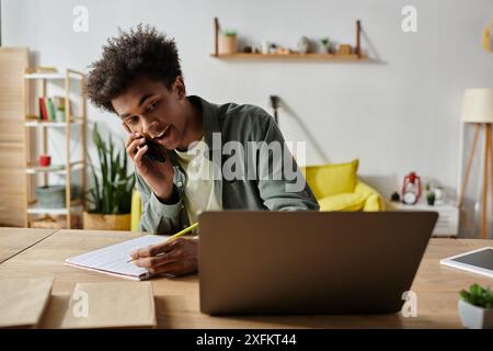 Un giovane afroamericano che parla al telefono, lavora al portatile e prende appunti alla scrivania. Foto Stock