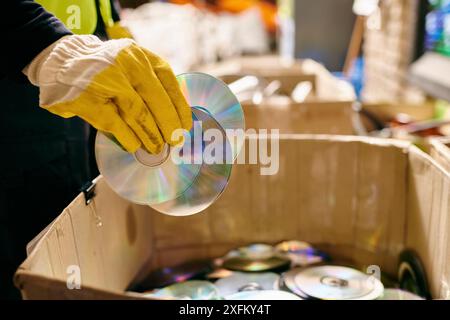 Una persona con guanti gialli tiene con cautela un CD all'interno di una scatola, smistando i rifiuti per un ambiente più pulito. Foto Stock