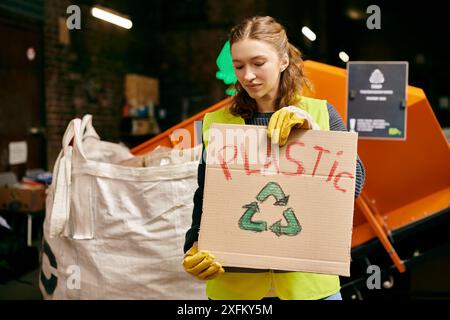 Un giovane volontario in guanti e giubbotto di sicurezza smista i rifiuti, tenendo un cartello con scritto plastica. Foto Stock