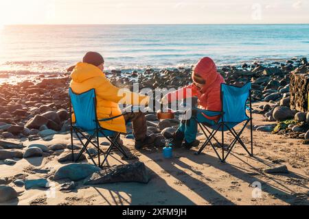 Due persone su sdraio insieme sulla spiaggia d'inverno, preparano tè caldo, caffè nella giornata del cielo azzurro e il mare sullo sfondo. Freddo inverno britannico. Vita Foto Stock