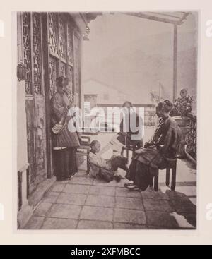 Foto d'epoca della vita quotidiana a Hong Kong del XIX secolo. Dopo cena. Illustrations of Qing China, 1875 questo è un incontro dopo cena sulla veranda di una casa di un cinese. L'intero circolo domestico fuma tabacco; ma le loro pipe differiscono dalle nostre. La vecchia donna e sua figlia usano un tubo che assomiglia alla "narghilè" con un piccolo compartimento pieno d'acqua, per raffreddare e purificare il fumo. Il paterfamilias si sta affondando e succhiando la fine di quello che sembra essere un bastone da passeggio molto formidabile ; ma in realtà è la sua pipa preferita, avendo una storia molto cara a Foto Stock