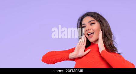 Ritratto di una giovane donna bruna allegra sorridente e gioiosa in camicia arancione che chiama qualcuno isolato su uno sfondo viola con spazio di copia Foto Stock