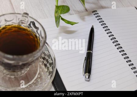 Una penna e una tazza di caffè si trovano su un notebook. Il caffè è caldo e la tazza viene riempita fino al bordo. La scena è semplice e tranquilla, con il caffè Foto Stock