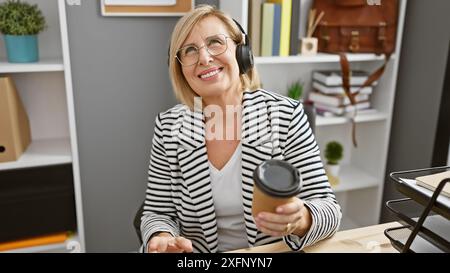 Una donna matura sorridente con le cuffie in un blazer a righe tiene il caffè in un ufficio moderno. Foto Stock