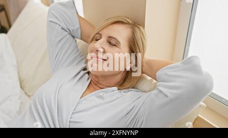 Una donna bionda di mezza età si rilassa nella sua camera da letto, incarnando il piacere e la tranquillità con un sorriso. Foto Stock