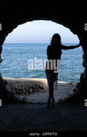 Questa immagine cattura la silhouette di una donna che si trova in piedi su una porta ad arco o in un'apertura, che sembra essere una grotta o un tunnel che conduce a una spiaggia Foto Stock