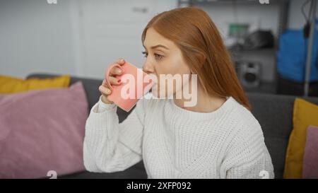 Una giovane donna rossa si gode una tazza di tè in un accogliente e moderno soggiorno dell'appartamento. Foto Stock