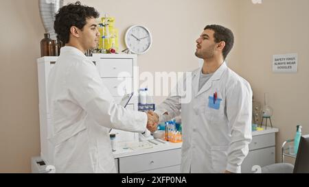 Due uomini in camici da laboratorio che stringono la mano in un laboratorio con attrezzature scientifiche e un cartello di sicurezza. Foto Stock