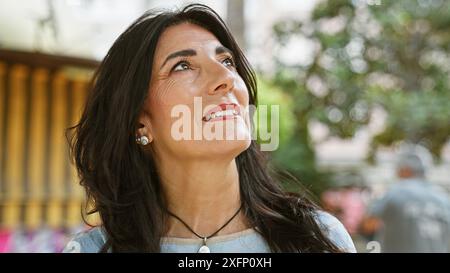 Una donna anziana ispanica contemplativa gode di un momento sereno in un giardino lussureggiante, evidenziando la sua maturità e gioia. Foto Stock