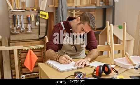 Uomo ispanico che lavora in multitasking, parla al telefono mentre disegna in un laboratorio di falegnameria Foto Stock