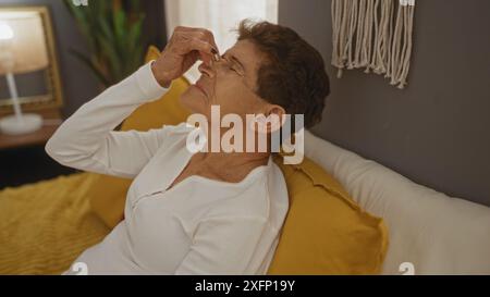 Un'anziana donna ispanica con i capelli corti si siede su un letto in una camera da letto, toccando il viso in un ambiente domestico. Foto Stock