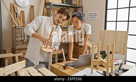 Due falegnami, un uomo e una donna, che lavorano insieme in un negozio di legno ben illuminato, discutono di un progetto con un portatile sul banco da lavoro Foto Stock