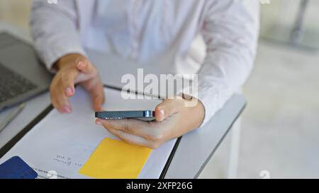 Una giovane donna d'affari ispanica che usa uno smartphone in un ufficio moderno, mentre documenti e una carta di credito sono sul tavolo. Foto Stock