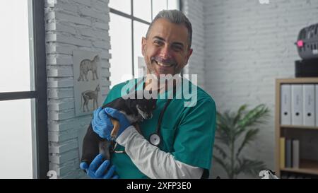 Veterinario maschio di mezza età che tiene un cane chihuahua in una stanza clinica, sorride alla telecamera, indossa uno stetoscopio e guanti Foto Stock