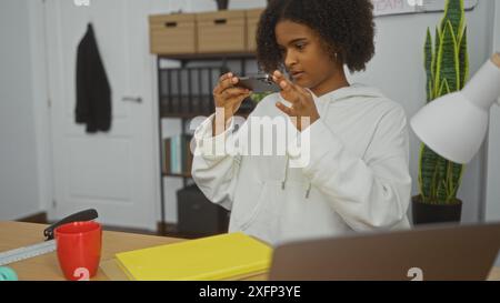 Una donna afroamericana in un ufficio che scatta una foto con il suo telefono, circondata da piante, cartelle e oggetti essenziali per il lavoro. Foto Stock