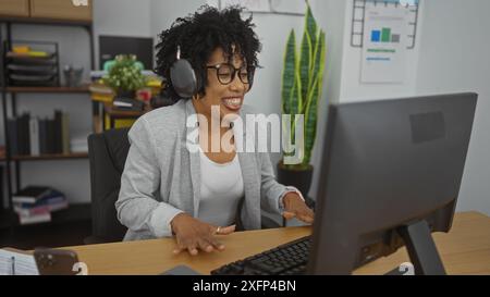 Una donna afroamericana sorridente siede in un ufficio con le cuffie e lavora al computer con i capelli ricci in un luogo di lavoro al chiuso. Foto Stock