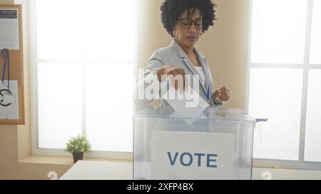 Una giovane donna afroamericana getta il suo voto in una sala elettorale del college con un ballottaggio in un ambiente al chiuso. Foto Stock