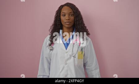 Una giovane donna afroamericana con i capelli ricci, con un camice da laboratorio bianco, si erge con sicurezza su uno sfondo rosa, mostrando il suo professionista in Foto Stock