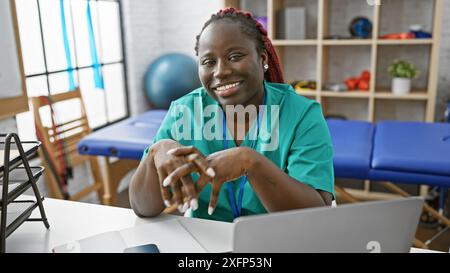 Donna afroamericana con trecce che sorride in una stanza clinica mentre si siede al suo posto di lavoro. Foto Stock